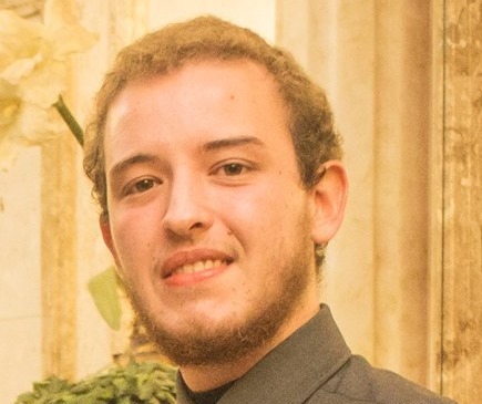 Fotografia colorida de Matheo Bernardino. Matheo está centralizado, tem cabelo encaracolado e loiro, é branco, está com uma camisa verde musgo e sorri para a foto. Matheo usa barba e tem olhos verdes. No fundo, uma parede amarelada e um arbusto na parte inferior esquerda.