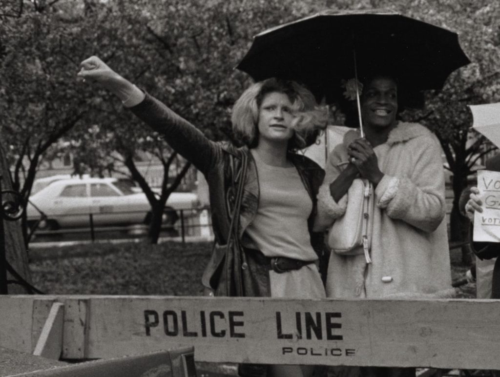 Fotografia de duas mulheres travestis, Sylvia Rivera e Marsha P. Johnson, porto-riquenha e negra, respectivamente. Na foto em PB, elas estão debaixo de um guarda-chuva, há uma cancela escrito "Police Line", Marsha está sorrindo, vestindo um casaco e Sylvia olha para a frente, com o punho fechado e lavantado. Ao fundo há algumas árvores e carros.