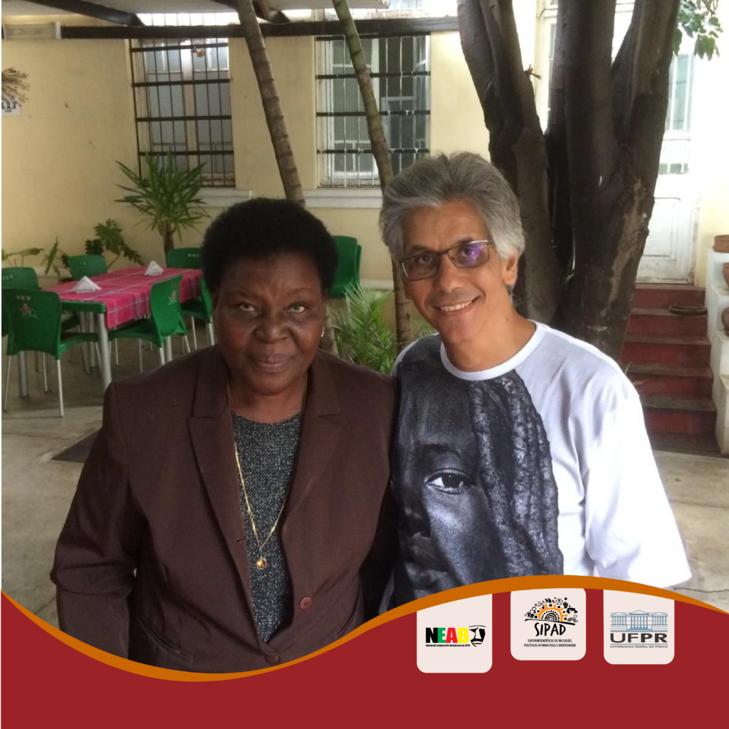 Foto da escritora Paulina Chiziane e do Professor Doutor Paulo Baptista da Silva, em encontro na Associação dos Escritores Moçambicanos, em Maputo, Moçambique. Paulina é uma mulher negra de cabelo curto cacheado, que no momento da foto usava um casaco marrom, uma blusa cinza e um colar. O professor Paulo é um homem negro de cabelos grisalhos curtos, que no momento da foto usava uma camiseta branca, cuja estampa é uma foto com filtro preto e branco de uma moça negra, com cabelo trançado. No fundo da foto encontra-se uma mesa com cadeiras verdes; uma parede de amarelo bem claro, com janelas e uma porta, a qual é acessada por uma escada de 5 degraus; e, logo atrás da escritora e do professor, têm-se alguns galhos de árvores. No rodapé a foto, é cortada por uma linha ondulada. Abaixo dessa linha, em fundo vermelho, encontram-se as logos do NEAB, da SIPAD e da UFPR.   