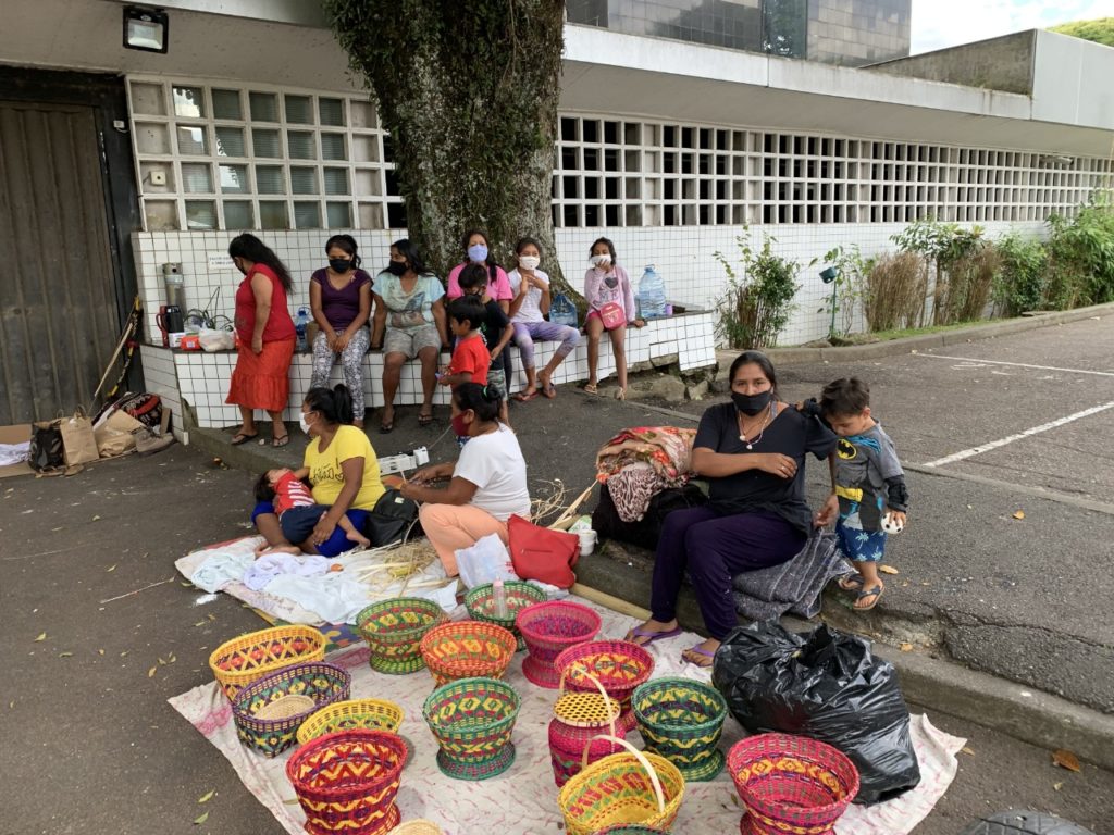 Imagem de uma família indígena (6 mulheres e 7 crianças em variadas idades e com as mesmas características físicas) expondo cestas artesanais coloridas no asfalto de uma rua. Atrás do grupo, está o Palácio das Araucárias com sua respectiva arborização.