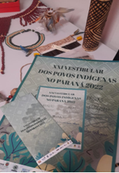 Sobre uma mesa branca, há vários objetos que representam diferentes povos indígenas. Em destaque há um cartaz, um folder e um panfleto sobrepostos. Os mesmos são na tonalidade verde, cinza e bege e divulgam o vigésimo segundo Vestibular Indígena. Neles estão contidas as seguintes informações: O Vestibular dos Povos Indígenas no Paraná será realizado nos dias 12 e 13 de junho de 2022. As inscrições começam no dia primeiro de abril e seguem até o último dia do mês. Para mais informações, acompanhe o site das universidades públicas paranaenses e as plataformas de notícias do Governo do Estado do Paraná. 