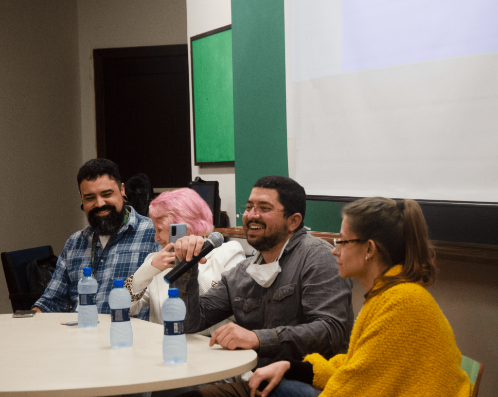 Na foto, tirada em um dos anfiteatros da Reitoria, há quatro pessoas sentadas atrás de uma mesa, frente a tela de projeção. Da esquerda para a direita, há um homem branco com cabelos curtos e barba, ele usa alargador e uma camisa xadrez azul. Ao seu lado, há uma mulher branca com os cabelos pintados de rosa, ela usa uma blusa branca e olha sorridente para o lado. Na sequência, há um homem branco com cabelos curtos e pretos, ele usa óculos, a máscara abaixada e uma camisa cinza, sorri e segura um microfone. Por último, há uma mulher branca com cabelos lisos, castanhos e presos, ela usa óculos e um casaco na cor mostarda. Na mesa, há algumas garrafas de água.
